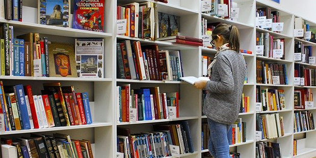 Bilden visar en tjej som står i ett bibliotek framför en bokhylla. 