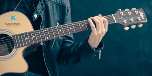 Bilden visar en person på nära håll som spelar gitarr och tar ett ackord. 