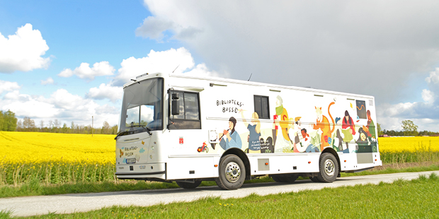 Bilden visar en bild på biblioteksbussen parkerad på en väg och en gul äng i bakgrunden. Bussen är vit i grunden och har illustrationer i färg på utsidan. 