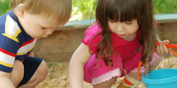 Bilden visar två barn som leker i en sandlåda. Barnet till vänster är en pojke som har en randig tröja och mörka shorts på sig. Barnet till höger är en flicka med långt hår och lugg, som har rosa tröja och rosa t-shirt på sig.