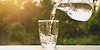 Ett glas fylls med vatten från en tillbringare, i sommarsol. 